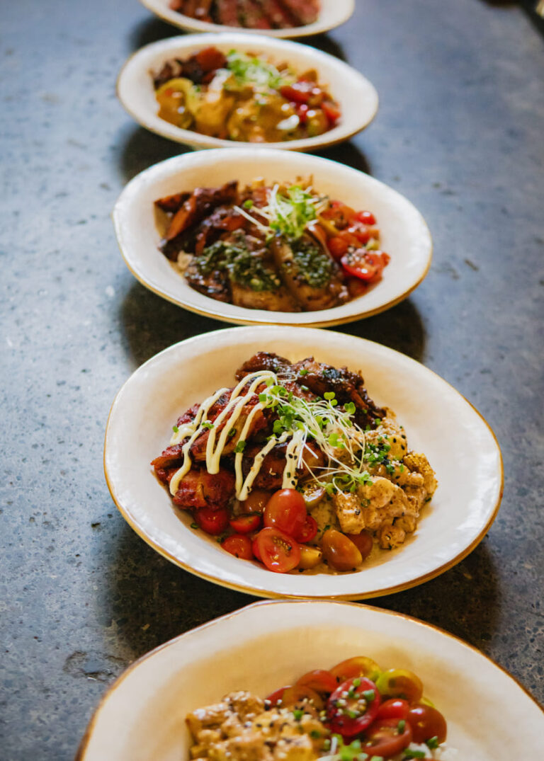 A row of white ceramic bowls with various dishes are arranged neatly on a dark countertop.