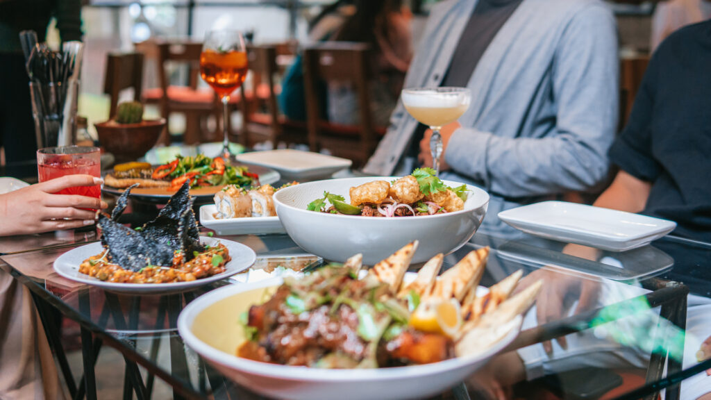 A group of people seated around a table filled with diverse dishes, including a bowl of salad, a plate of grilled meat with breadsticks, a dish with black seaweed, and various drinks.