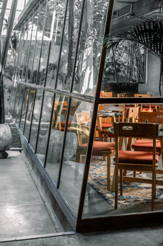 A modern glass-walled cafe with wooden tables and chairs, plants in large pots, and natural light streaming in from outside.