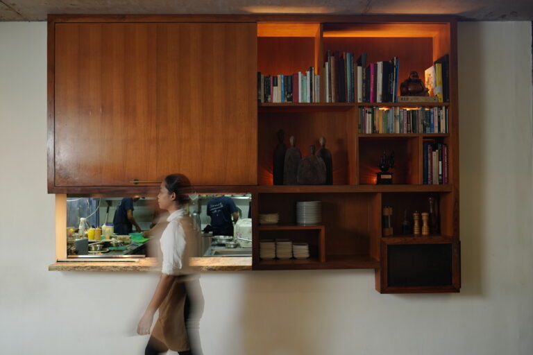 A woman strolls by a bookcase filled with books in a cozy kitchen setting.