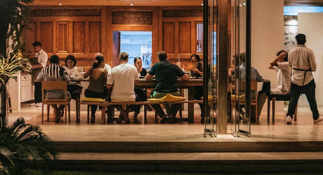 Group dining experience in a modern restaurant setting, featuring a long wooden table with seven diners engaged in conversation and enjoying their meals.