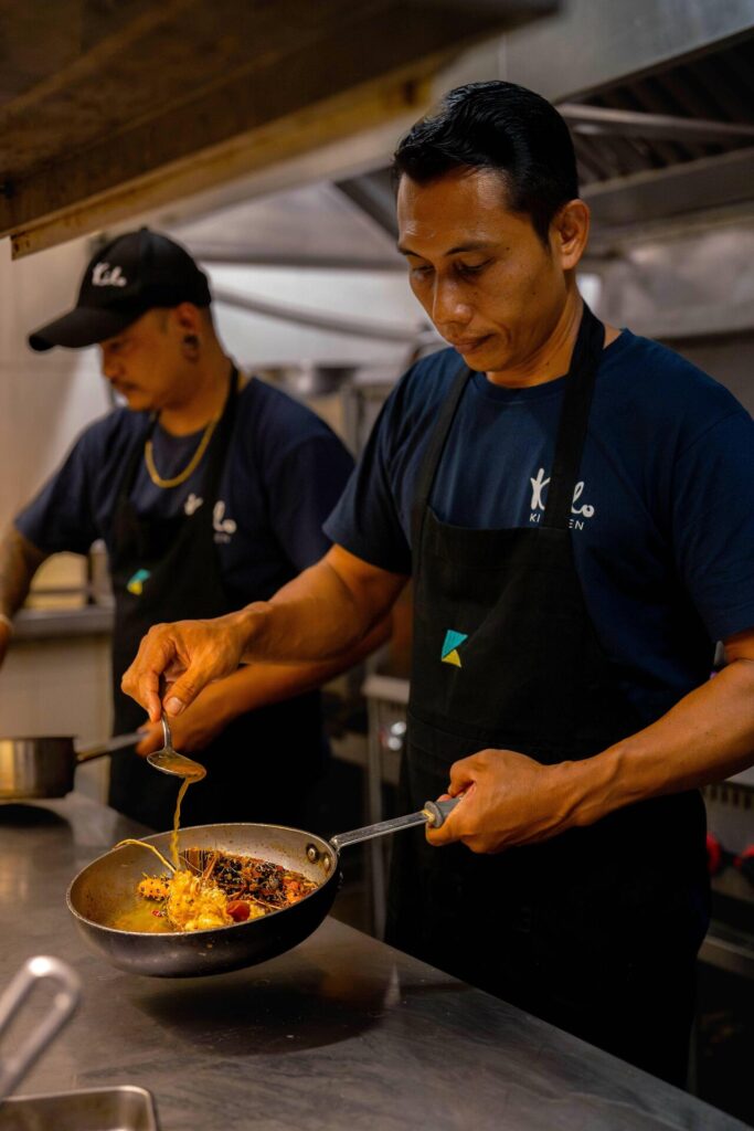 A chef in a dark apron skillfully pours sauce from a ladle into a pan of colorful, plated food in a bustling kitchen.