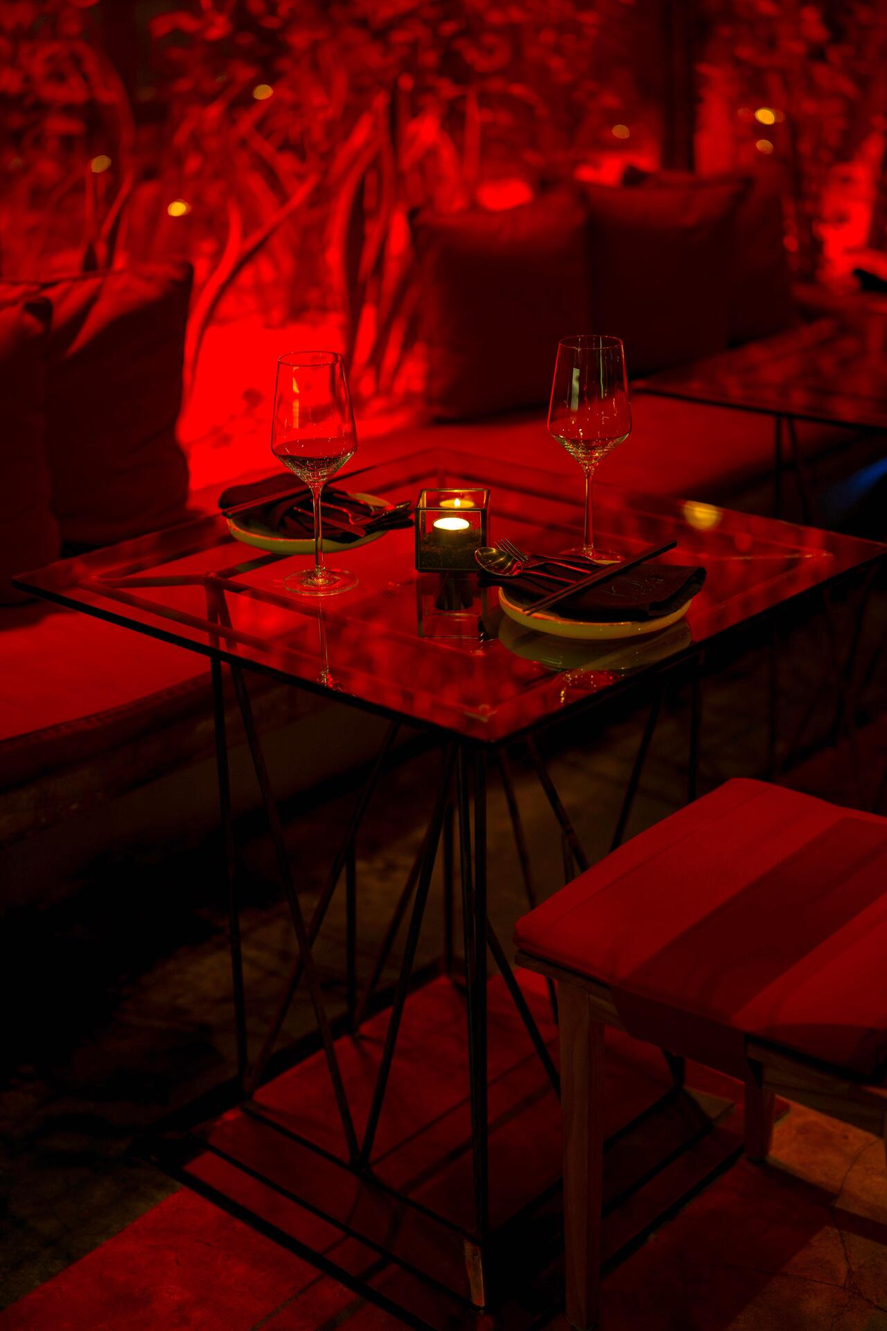Intimate dining setup with a glass table, wine glasses, and candles, surrounded by plush red cushions in a dimly lit atmosphere.