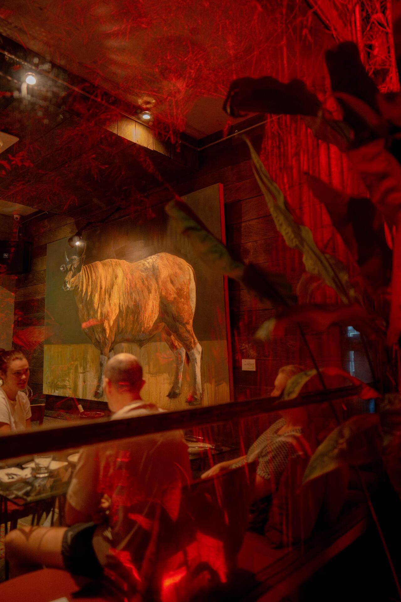 A cozy restaurant interior features a large, illuminated cow painting against a backdrop of warm red lighting and tropical plants.