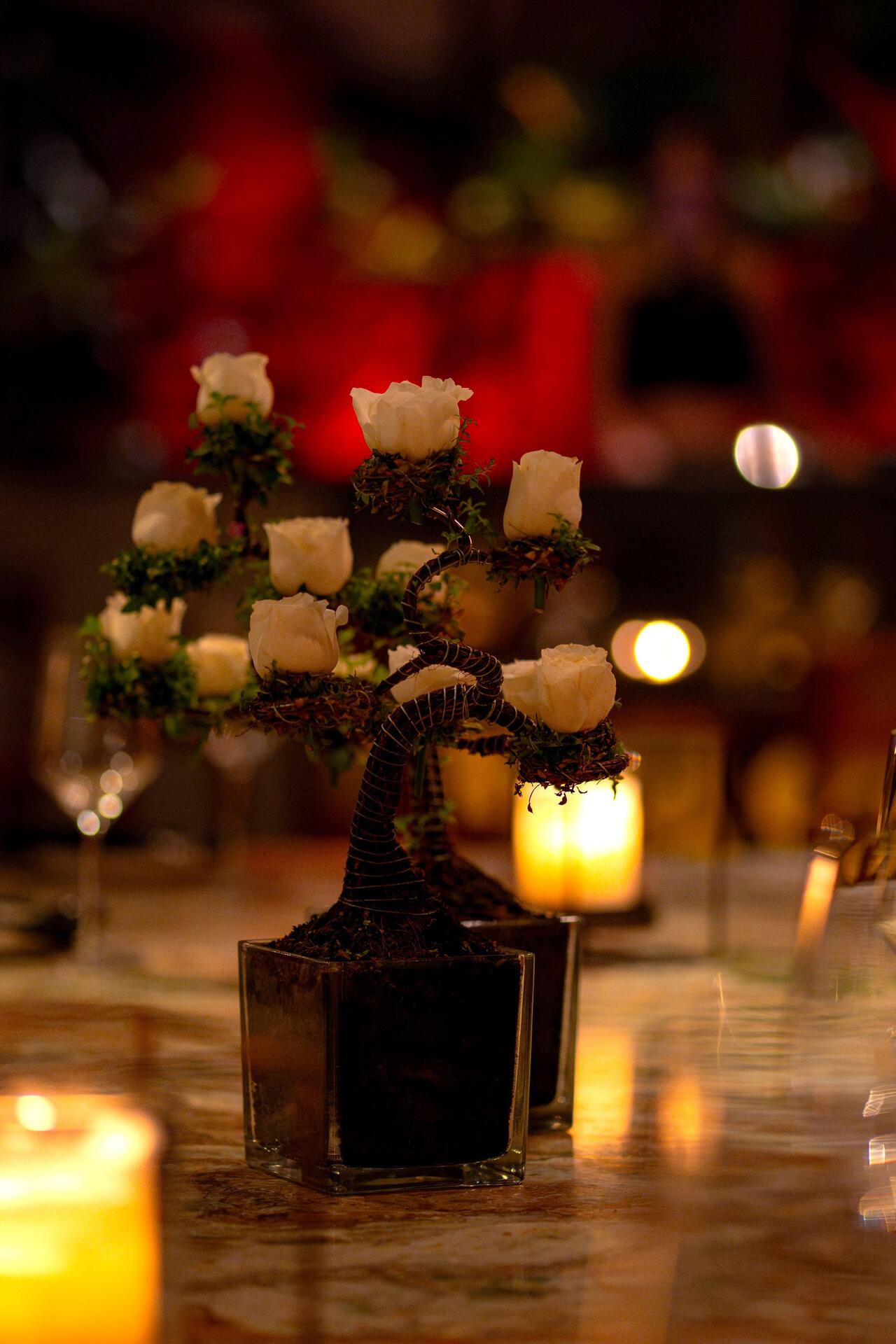 A small bonsai-style arrangement with white rosebuds in a glass pot, placed on a table with candles and blurred background lights.