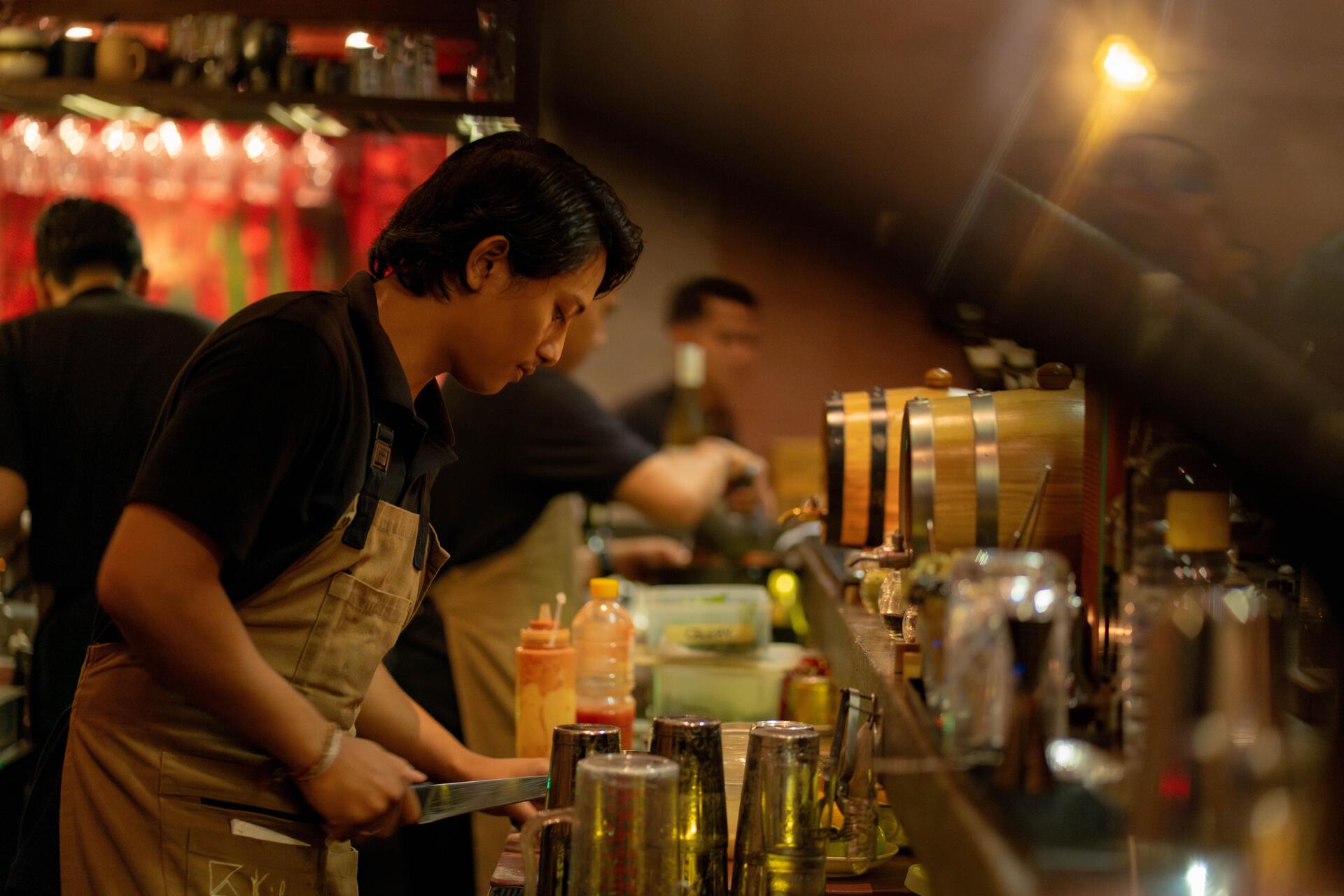 A bartender in an apron preparing drinks with various bottles and utensils on a busy bar counter, illuminated by warm lights.