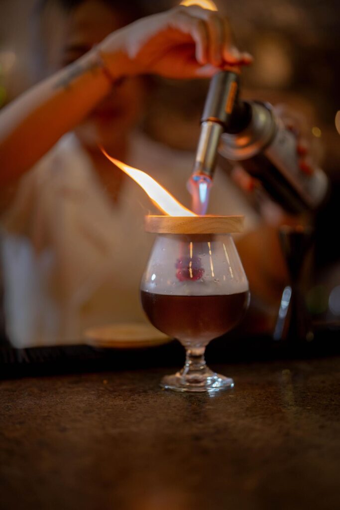 A bartender uses a torch to ignite a cocktail with a wooden lid, creating a striking flame atop a glass drink on a bar counter.