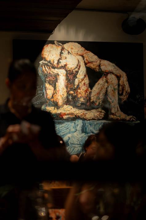 A blurred foreground shows people in a dimly lit room, with a large, abstract painting of a seated, contemplative figure displayed on the wall behind them.