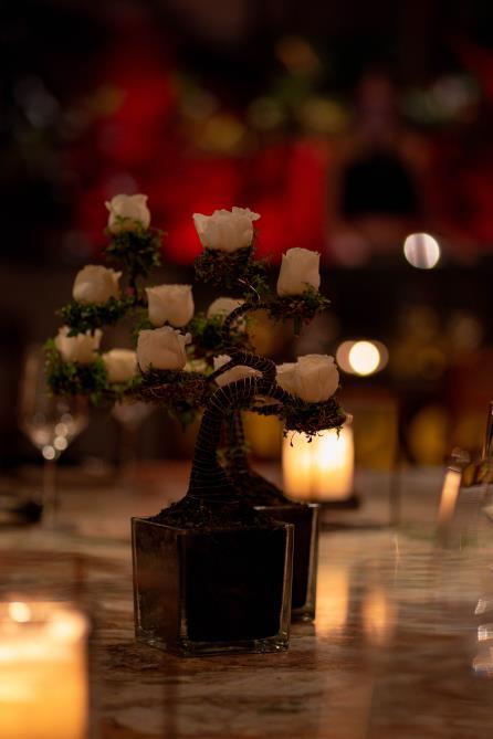 A small bonsai-style arrangement with white rosebuds in a glass pot, placed on a table with candles and blurred background lights.