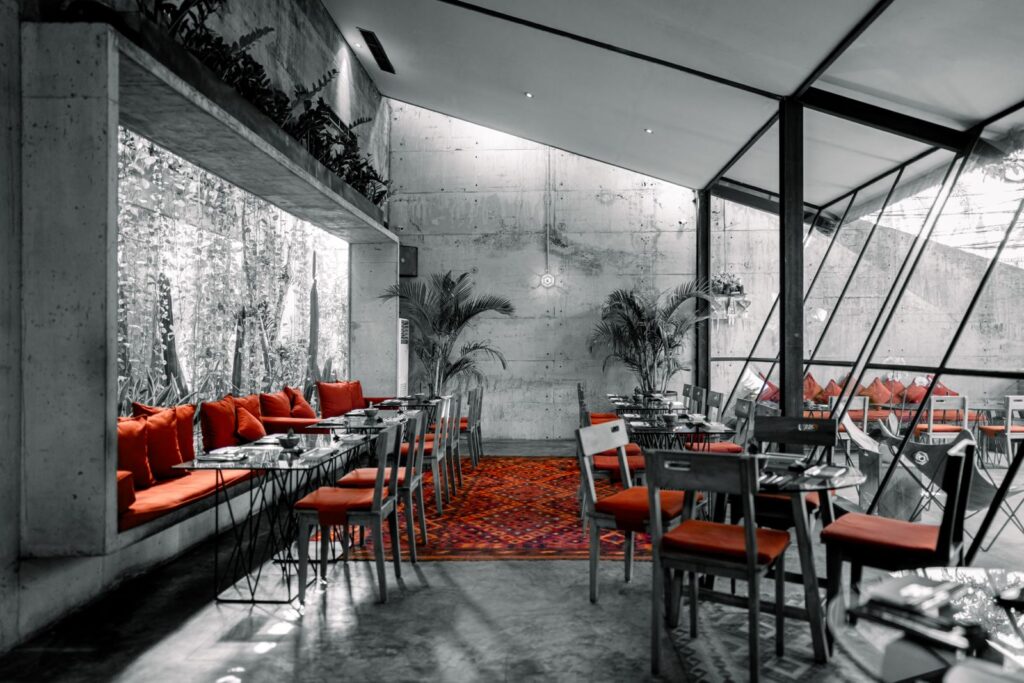 Modern restaurant interior with concrete walls, glass windows, red cushioned seating, and a colorful red rug on the floor.