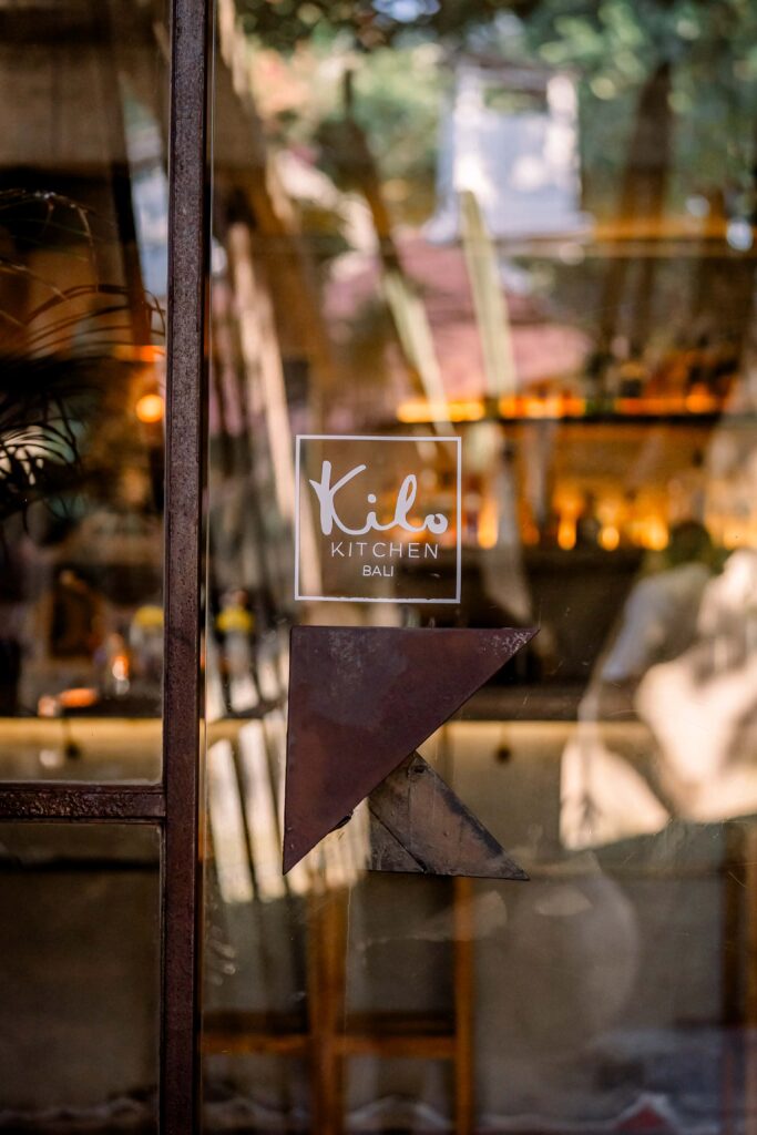 Kilo Kitchen Bali logo displayed on a glass panel with the restaurant's dining area in the background.