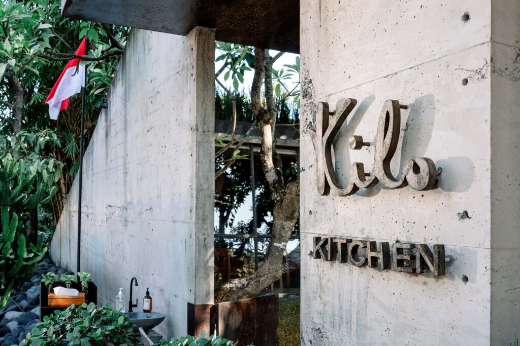 Exterior entrance of Kilo Kitchen Bali featuring its concrete facade, signature logo, and tropical greenery.