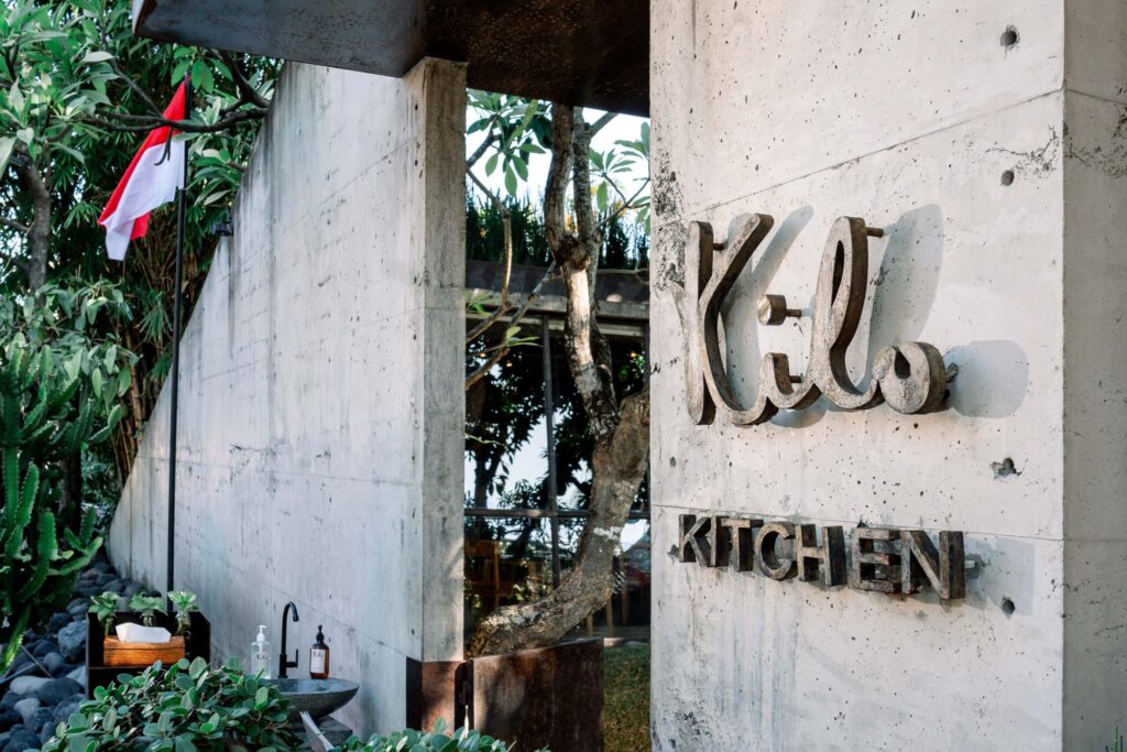 Exterior entrance of Kilo Kitchen Bali featuring its concrete facade, signature logo, and tropical greenery.