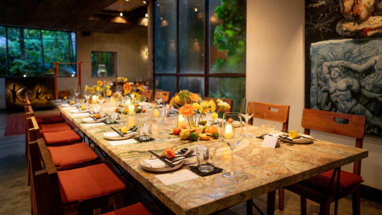 A long, beautifully decorated communal dining table at Kilo, set for a party with floral centerpieces, candles, and garden views.