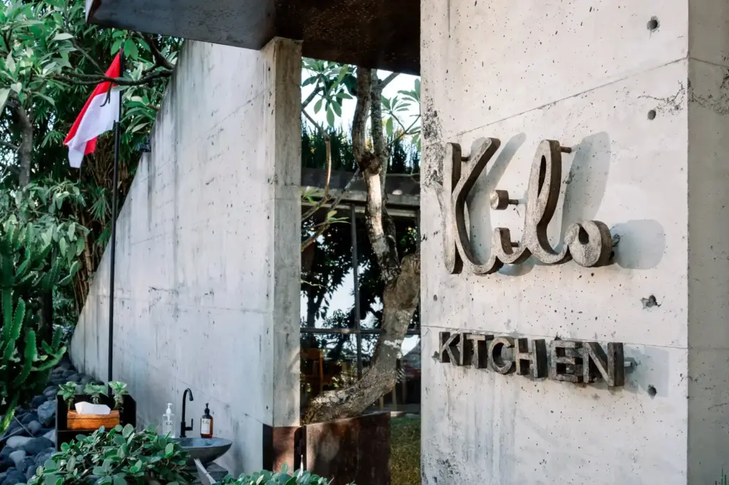 The exterior concrete entrance of Kilo Kitchen, featuring its logo mounted on the wall alongside lush greenery and an Indonesian flag