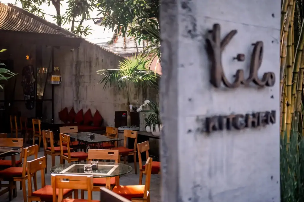 An outdoor patio dining area at Kilo Kitchen featuring wooden tables and chairs, surrounded by lush green foliage and tropical decor