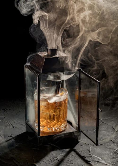 A whiskey glass sitting inside a glass lantern filled with dense smoke billowing out the top.