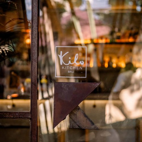 Kilo Kitchen Bali logo displayed on a glass panel with the restaurant's dining area in the background.