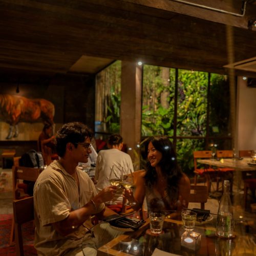 A couple enjoys drinks at a dimly lit restaurant with lush greenery visible through a window