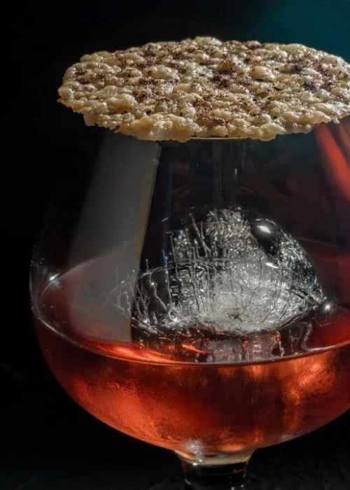 An amber cocktail with a single large ice sphere served in a snifter glass topped with a textured wafer.