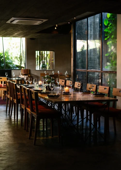 Cozy dining area at Kilo Kitchen featuring vibrant orange cushions, glass tables, and lush green plants behind large glass windows.